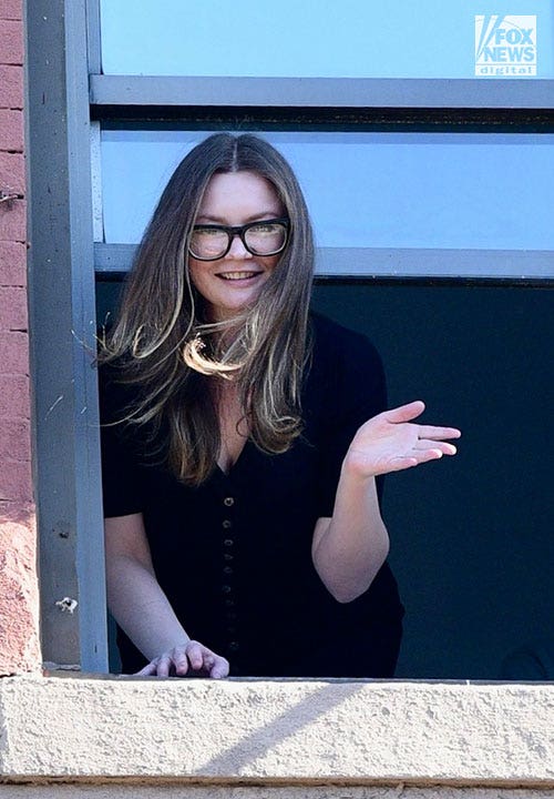 Anna Sorokin waves out the window of a brick building in a black top and glasses.