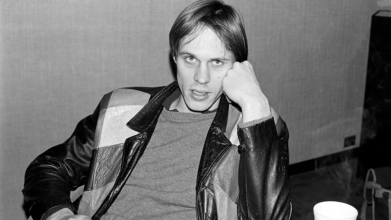 Tom Verlaine with his a fist to his face in a patterned leather jacket stares at the camera