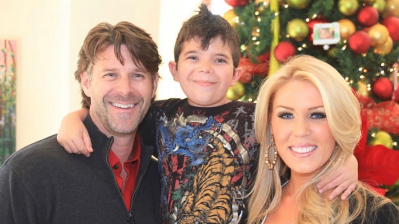 Slade Smiley in a black zip-up and red button down smiles for a photo next to son Grayson in a printed graphic shirt with arms wrapped around Slade and Christine Rossi, who is wearing a sparkly gold shirt and black sweater
