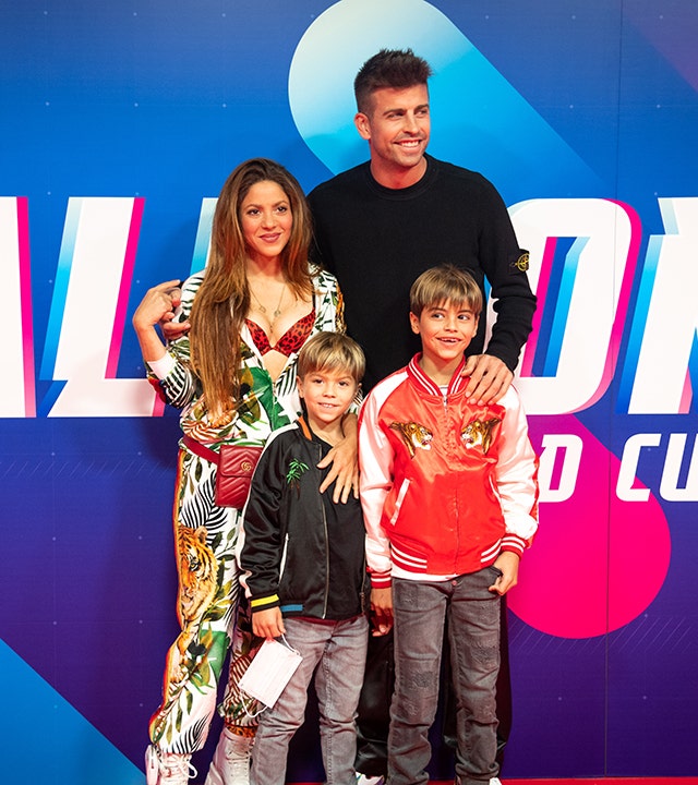 Shakira exposes her red leopard bra wearing a white outfit with jungle animals on it, has Gerard Piqué's arm wrapped around her, he is wearing black, and their two sons both in relaxed bomber jackets stand in front of them