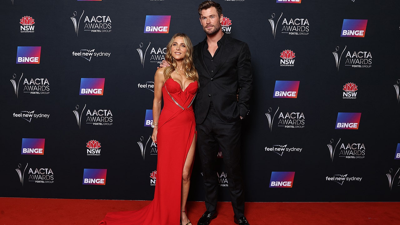 Chris Hemsworth in a black suit and jacket poses on the red carpet next to Elsa Pataky in a busty red gown in Sydney, Australia