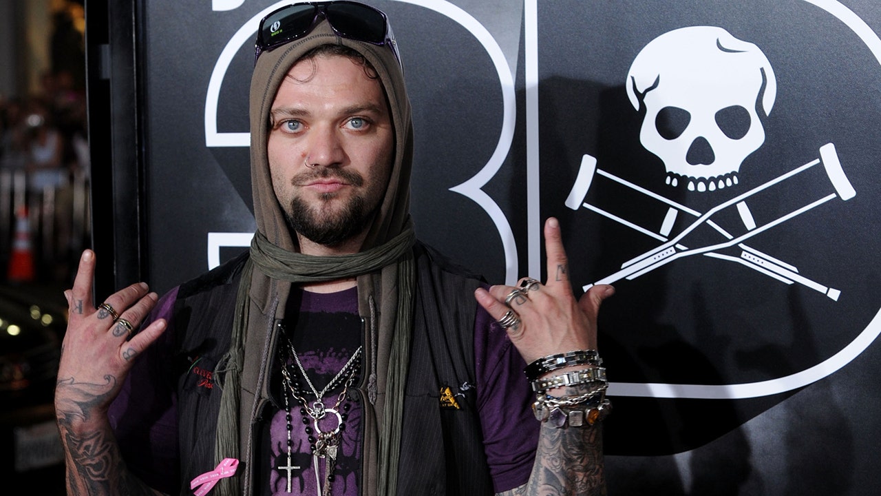 Bam Margera flashes his hands on the red carpet with sunglasses on his head and a scarf covering his head in a purple shirt