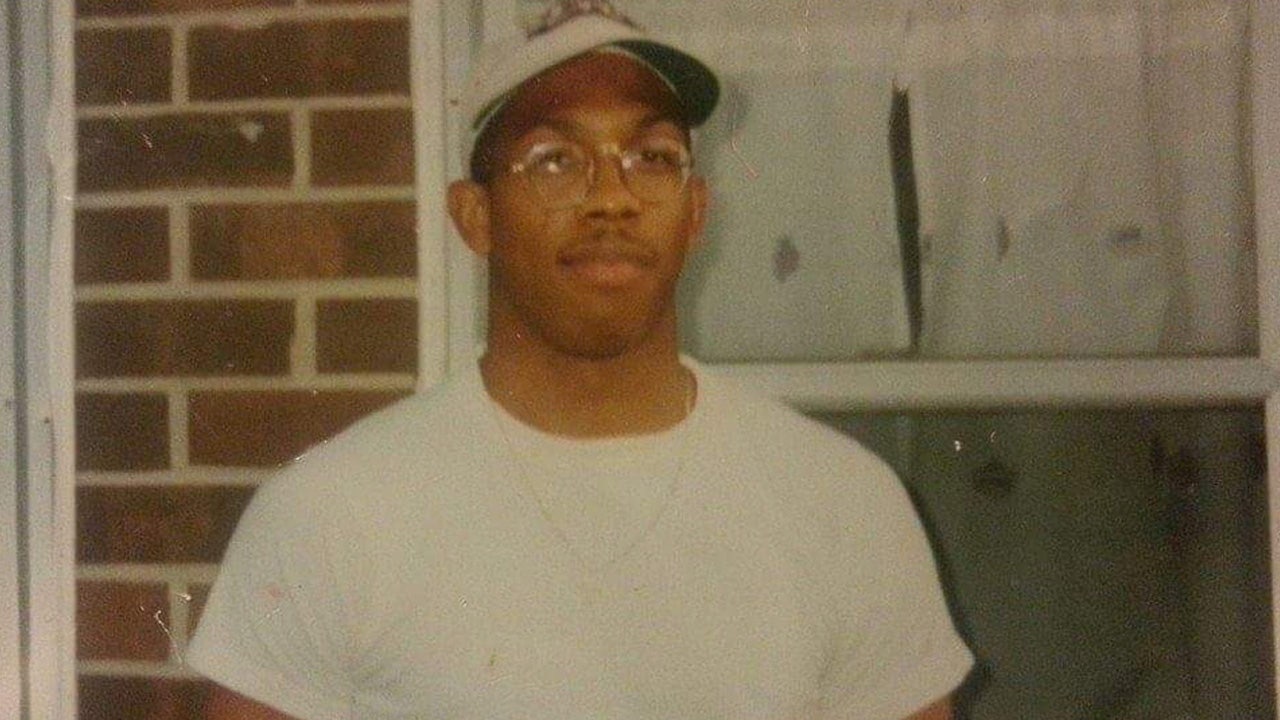 Damion Cooper wearing a white T-shirt and ball cap, standing in front of a brick home