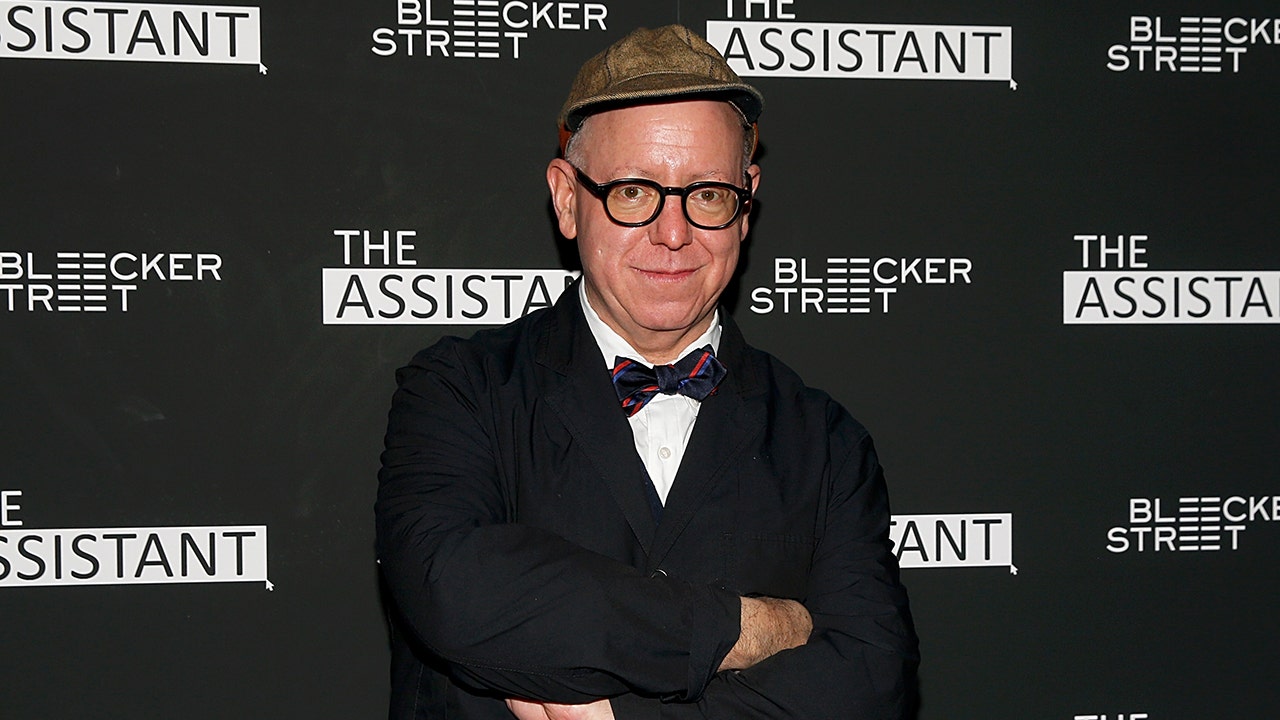 Writer James Schamus soft smiles for the camera at a screening of "The Assistant" wearing a suit and bow-tie, with his arms crossed, dark rimmed glasses and a hat