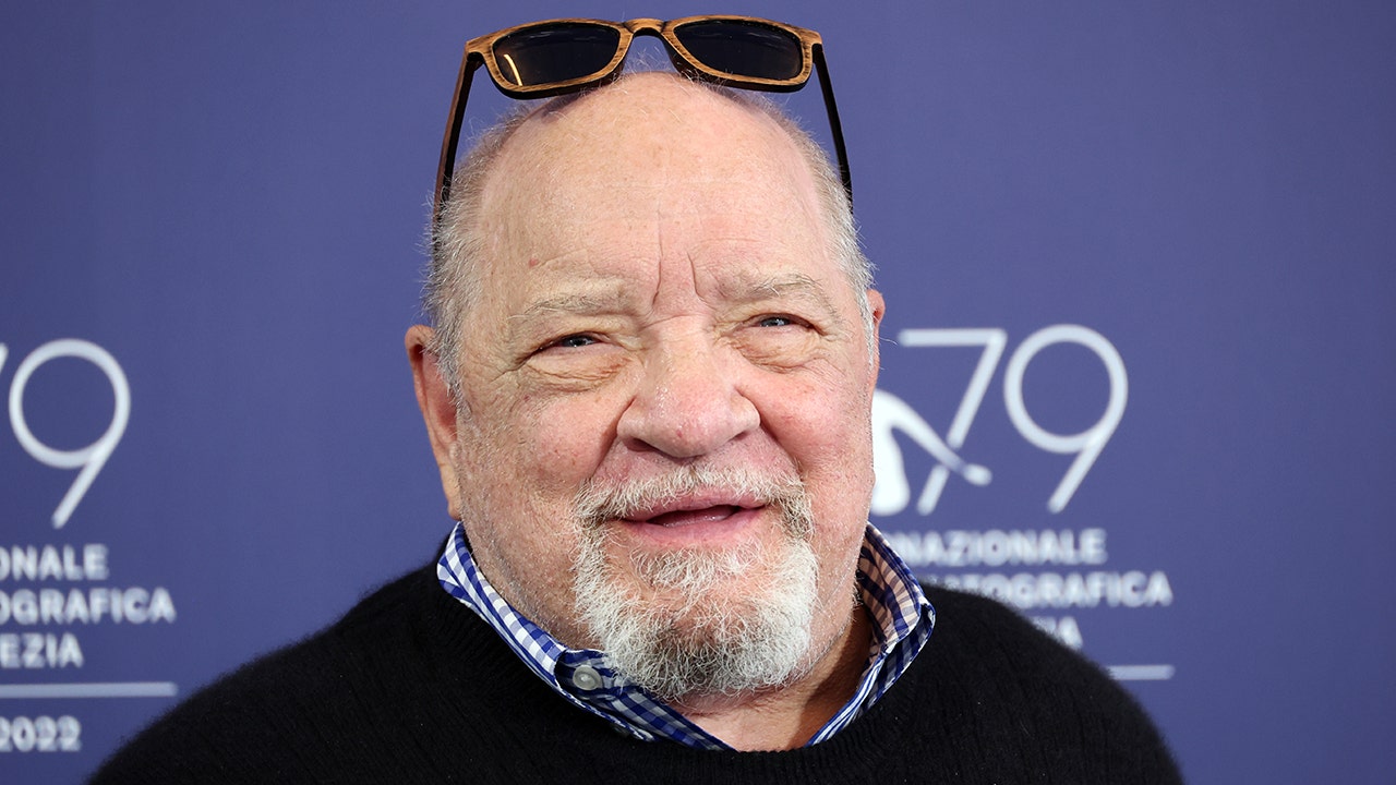 Paul Schrader smiles with his sunglasses on top of his head, wearing a checkered shirt and sweater over on the red carpet