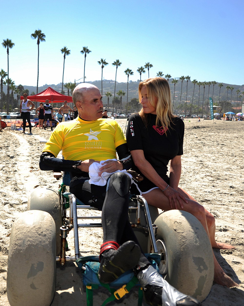 A disabled veteran wearing a yellow shirt in a wheelchair chatting with Bo Derek in a black outfit at the beach