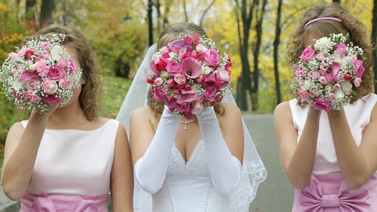 Bride poses with two bridesmaids in pink dresses while each of them hold a bouquet in front of their faces.