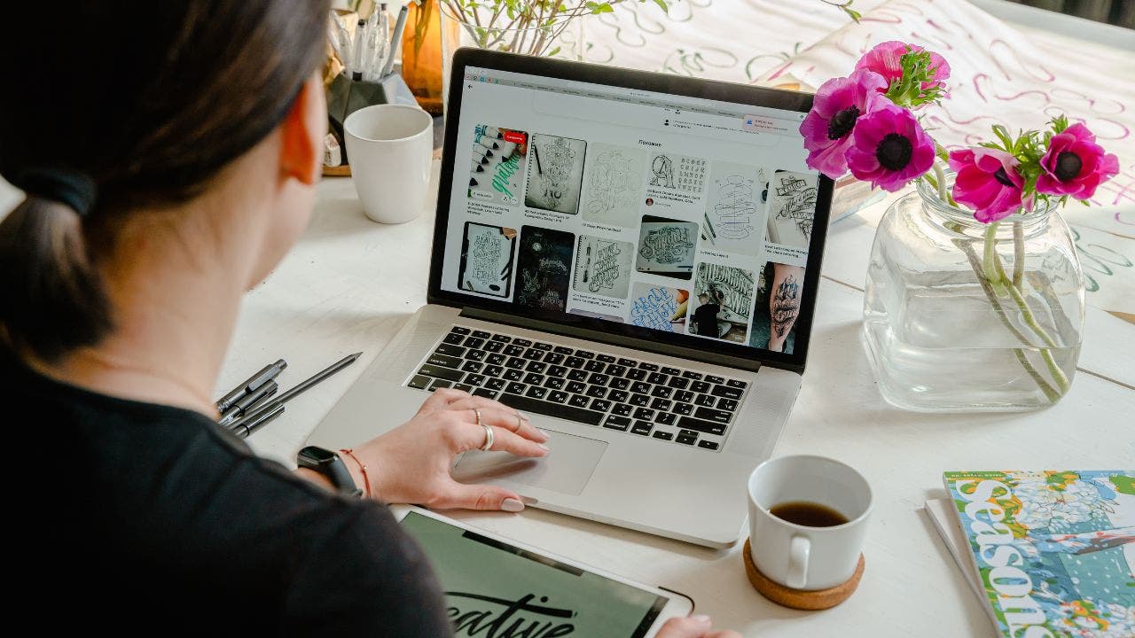 Woman looking down at laptop, pink daisies in vase, with a cup of coffee next to her on table