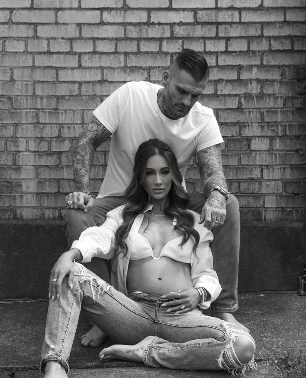 Leah Van Dale wearing a white bikini top with an open white blouse and sleeves sitting down with Matt Polinsky sitting above her wearing a white shirt and jeans in a black and white photo