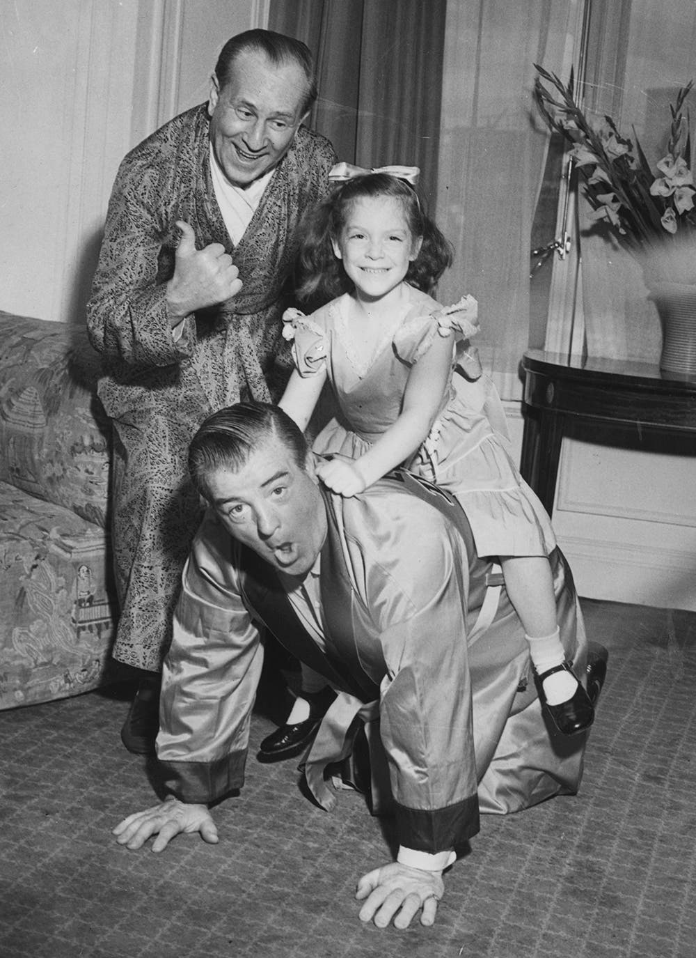 Abbott and Costello in suits and smiling at the camera as they play horse ride with daughter Chris Costello