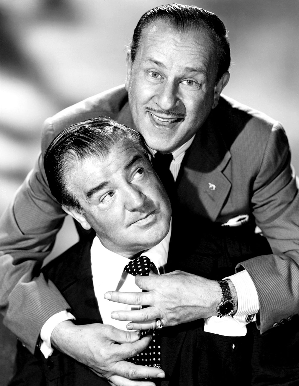 Bud Abbott in a suit embracing Lou Costello who is also in a suit and looking above at Lou Costello