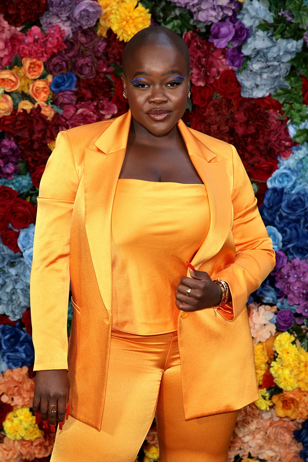 Achieng Agutu stands in front of a wall of flowers wearing a bright gold suit.
