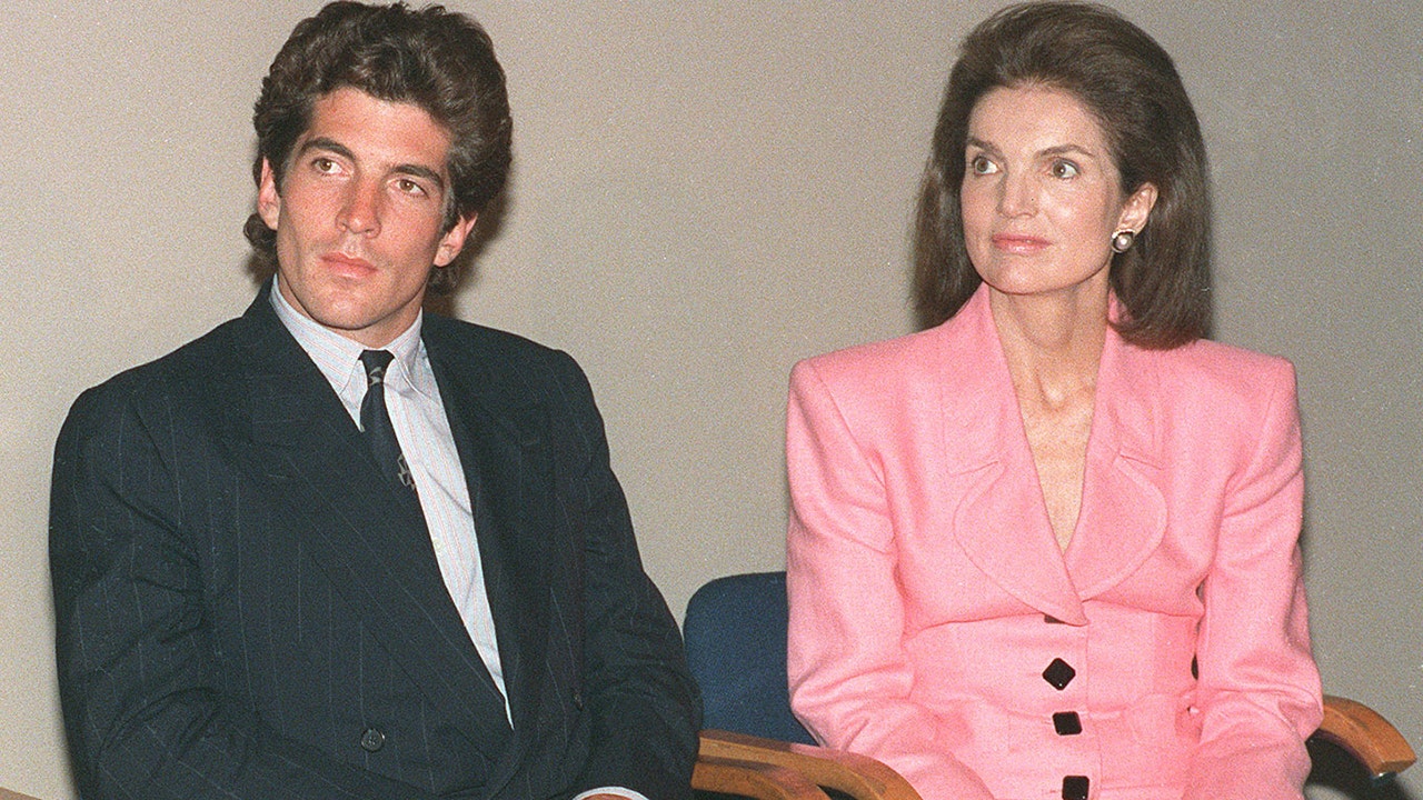 Jackie Kennedy, wearing a bright pink suit, sits with her son, John F. Kennedy Jr., in a suit and tie.
