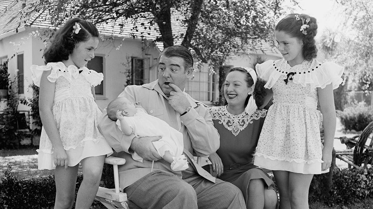 Lou Costello making a funny face as he holds his infant daughter with his older daughters and wife smiling in awe