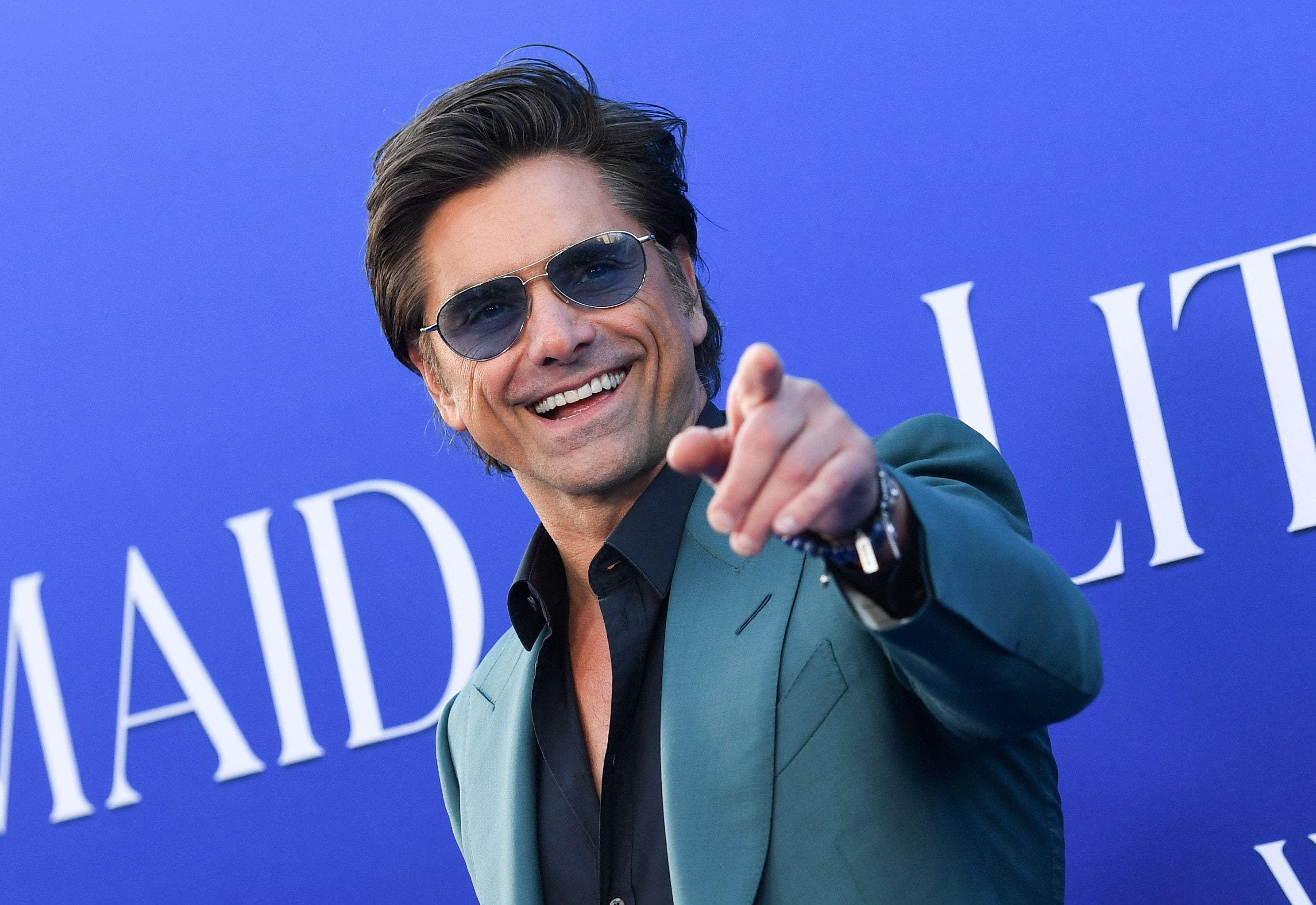 John Stamos smiling and wearing sunglasses and pointing