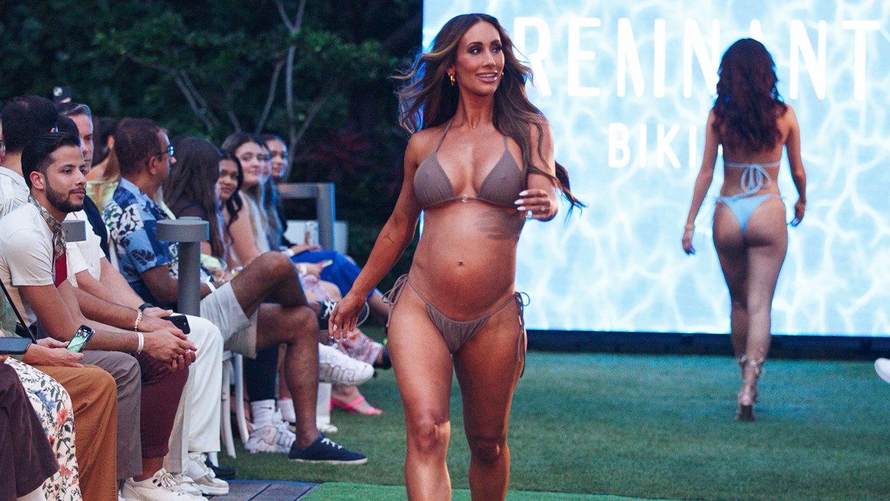 Leah Van Dale in a dark beige bikini walking down a green runway with a crowd on her left side