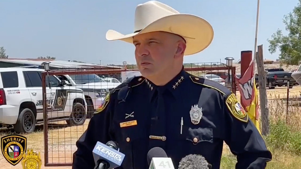 Man in cowboy hat stands in front of a police car talking to reporters.