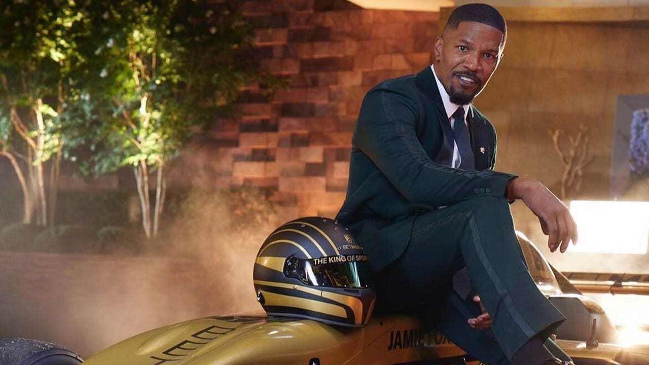 Jamie Foxx in a black suit sits on a sports car for a BTMG ad.