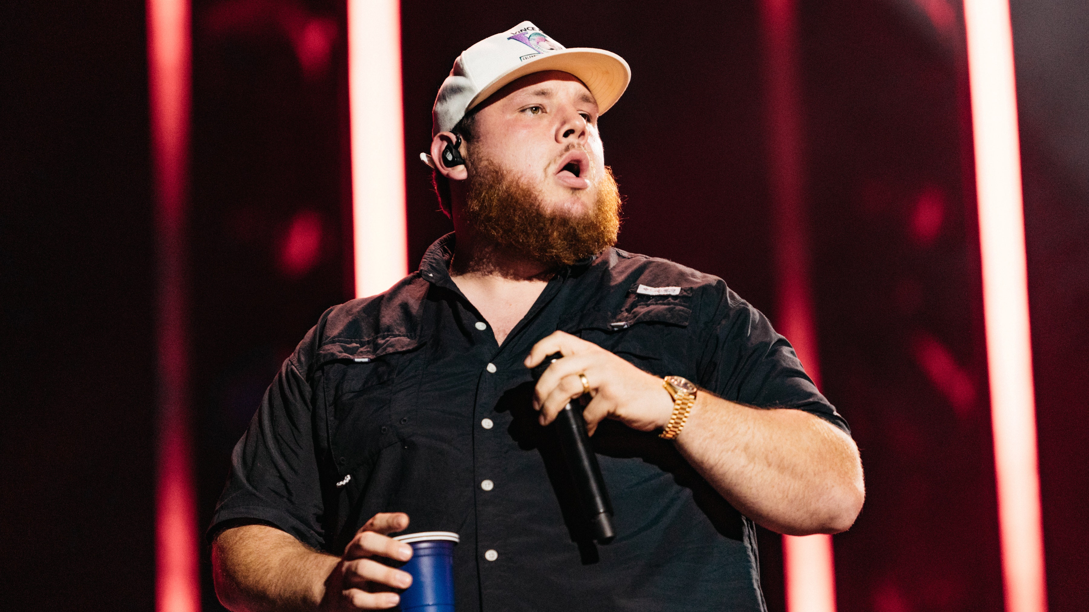 Luke Combs in a black shirt and light baseball cap holding a microphone and blue solo cup