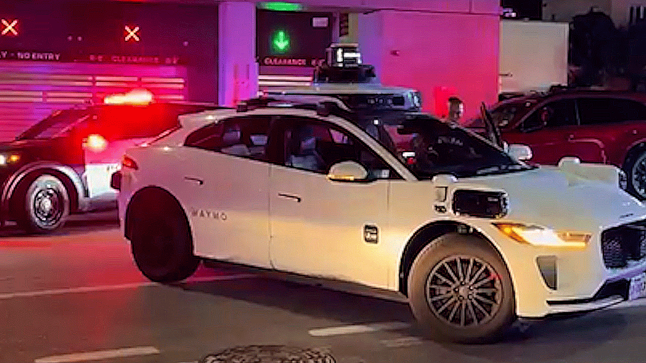 A police officer opens the driver-side door of a white Waymo autonomous SUV in downtown Austin at night, with patrol lights flashing nearby.