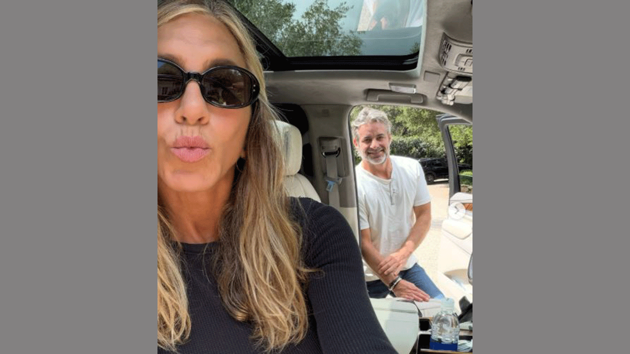 Jennifer Aniston blows a kiss in a selfie with JIm Curtis standing behind her